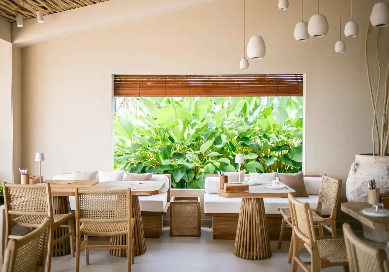A bright, modern cafe interior with natural wood furniture, a large window, and lush greenery.