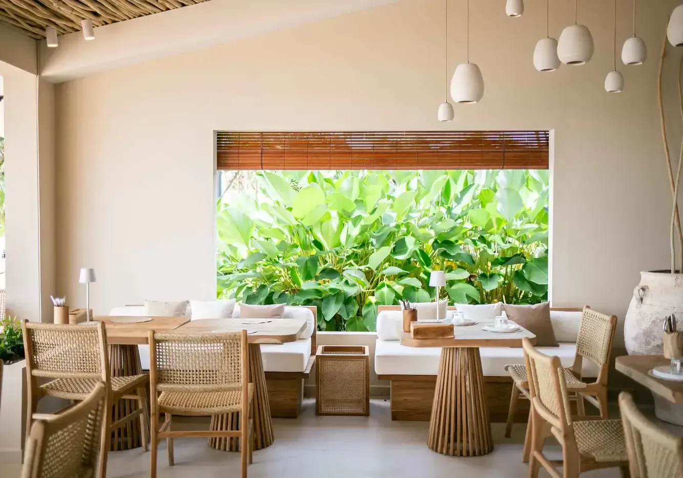 A bright, modern cafe interior with natural wood furniture, a large window, and lush greenery.